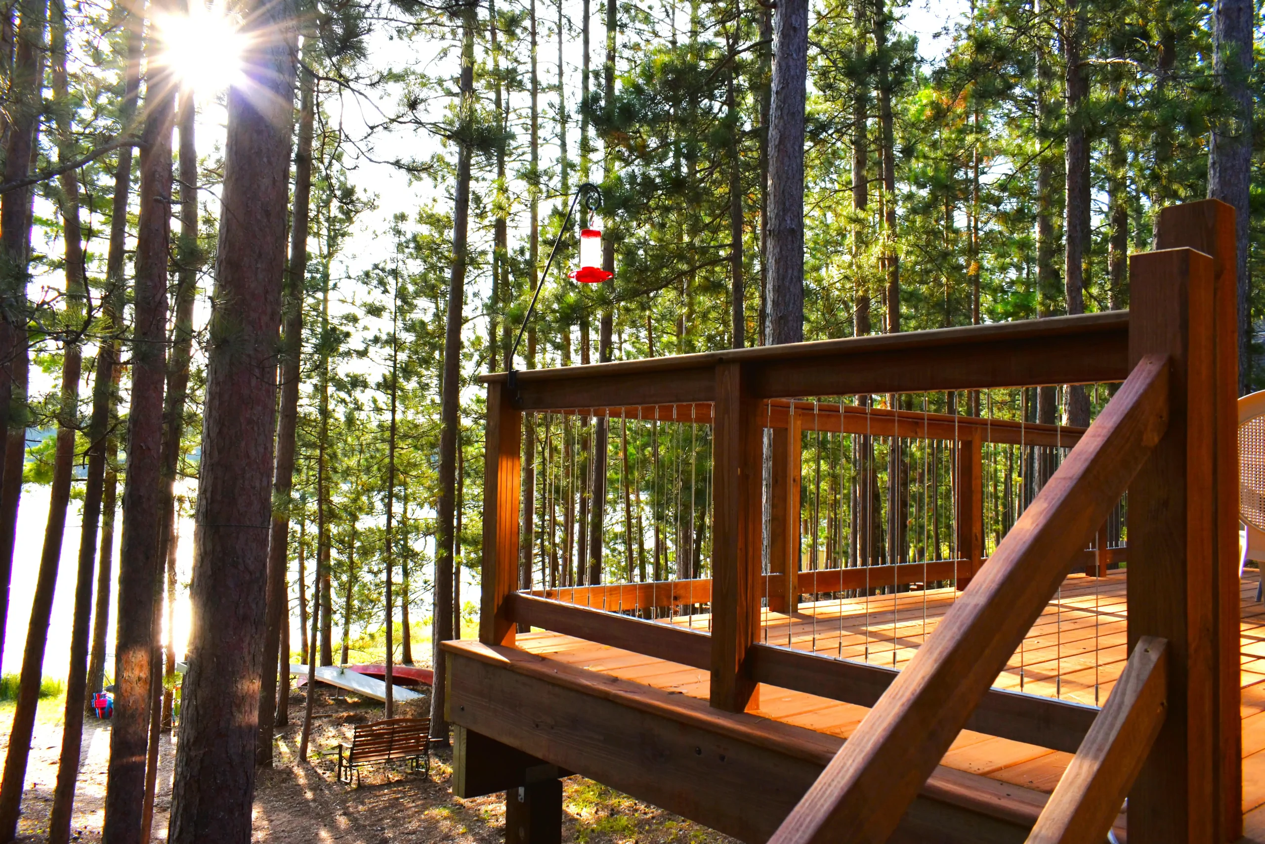 Wooden deck railing overlooking a serene lakeside view, surrounded by tall pine trees and a hummingbird feeder, capturing the essence of outdoor living and home improvement.
